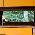 キッチンからリビングのFIX窓を通して借景を楽しむ オレンジ色のキッチンと渓流の木々の緑